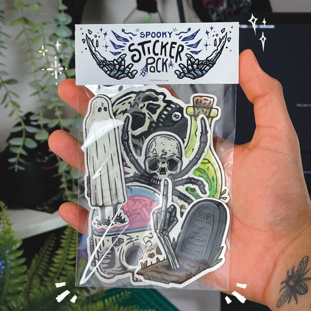 Hand holding a sticker pack with spooky-themed cartoon stickers, with a header saying "SPOOKY STICKER PACK" against a background of plants
