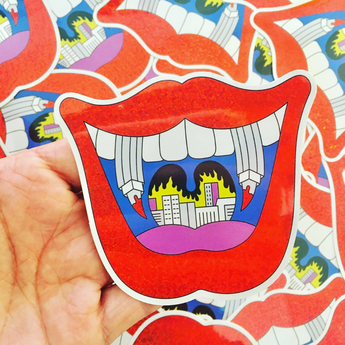 Hand holding a sticker of red lips with a cityscape and flames inside the mouth, created bu Jeune Woof.