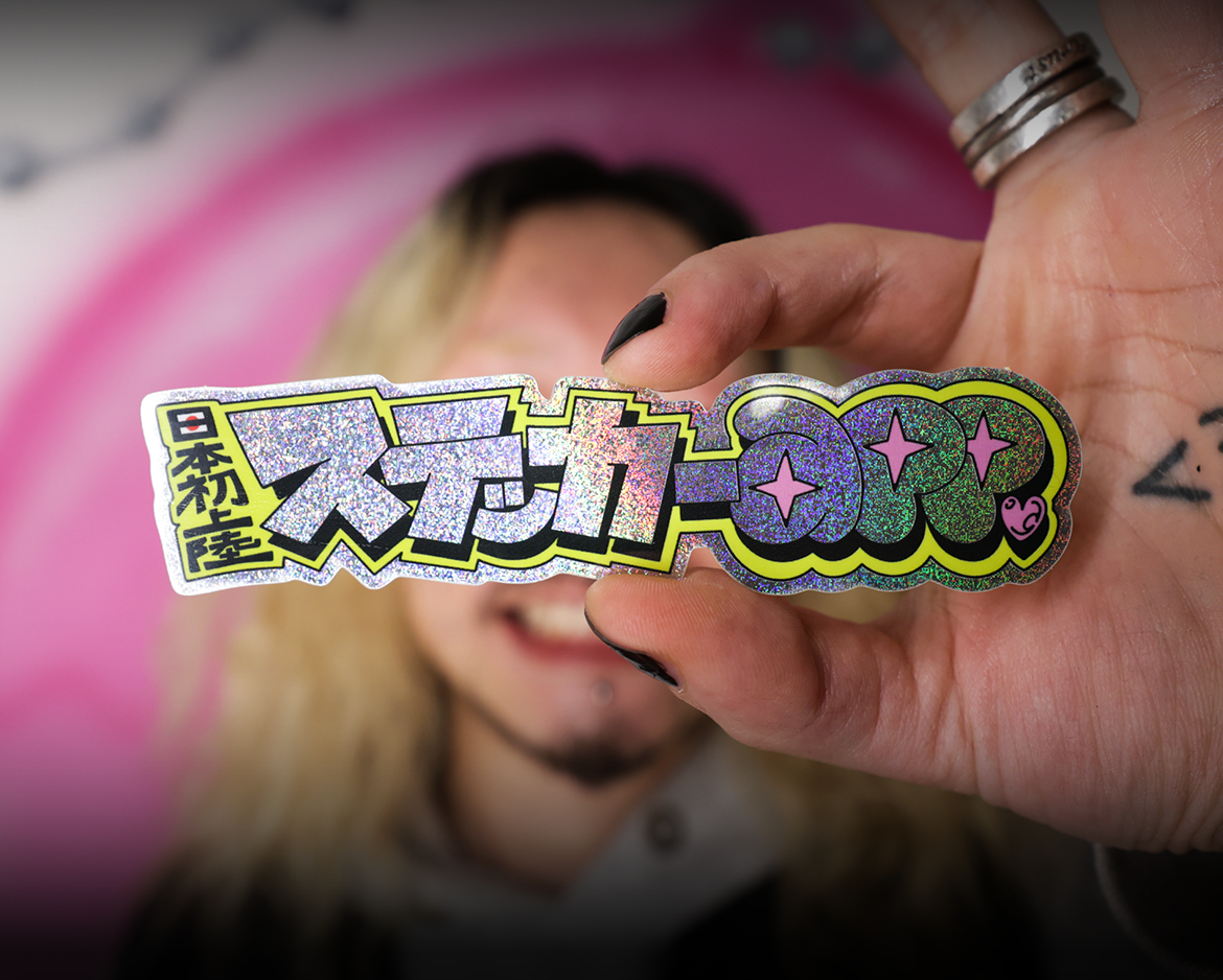 Close-up of a hand holding a holographic die-cut sticker featuring bold Japanese-inspired lettering