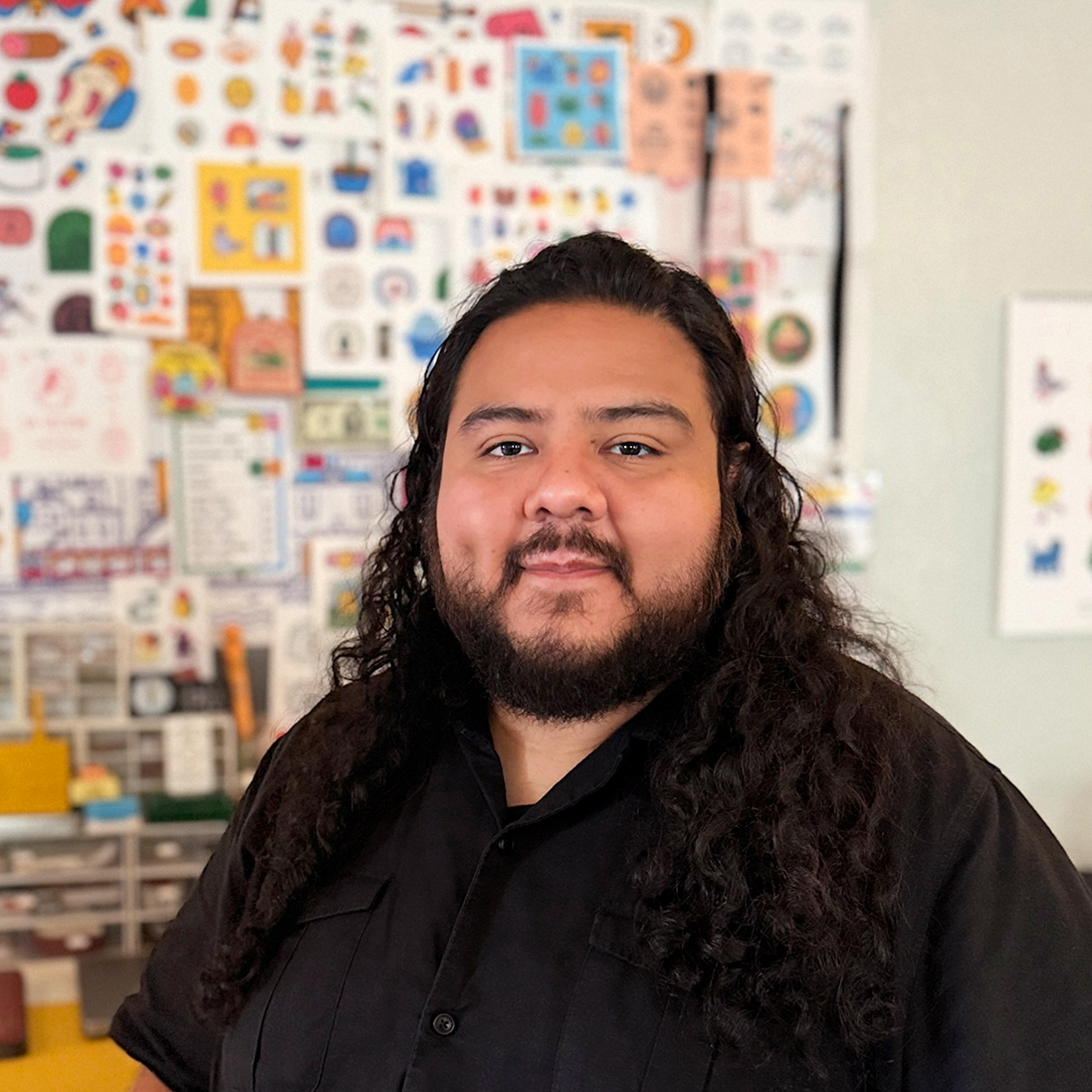 Damiel Orellana wearing a black shirt, stands in front of a colorful wall filled with various art prints, stickers and designs.