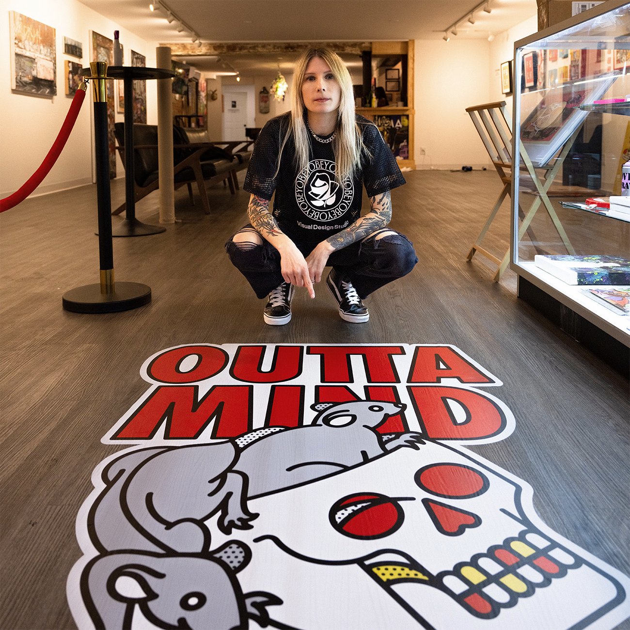 Person kneeling next to colorful "OUTTA MIND" sticker on a gallery floor, surrounded by artwork.