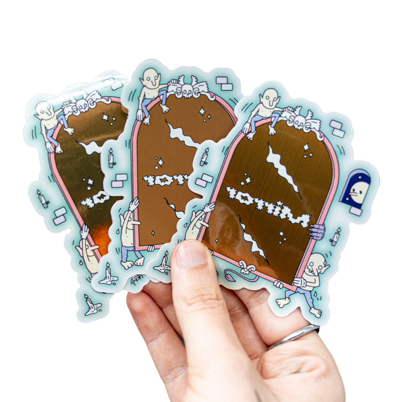 Hand holding three metallic stickers featuring cartoon skeletons climbing on bronze mirrors with playful designs.