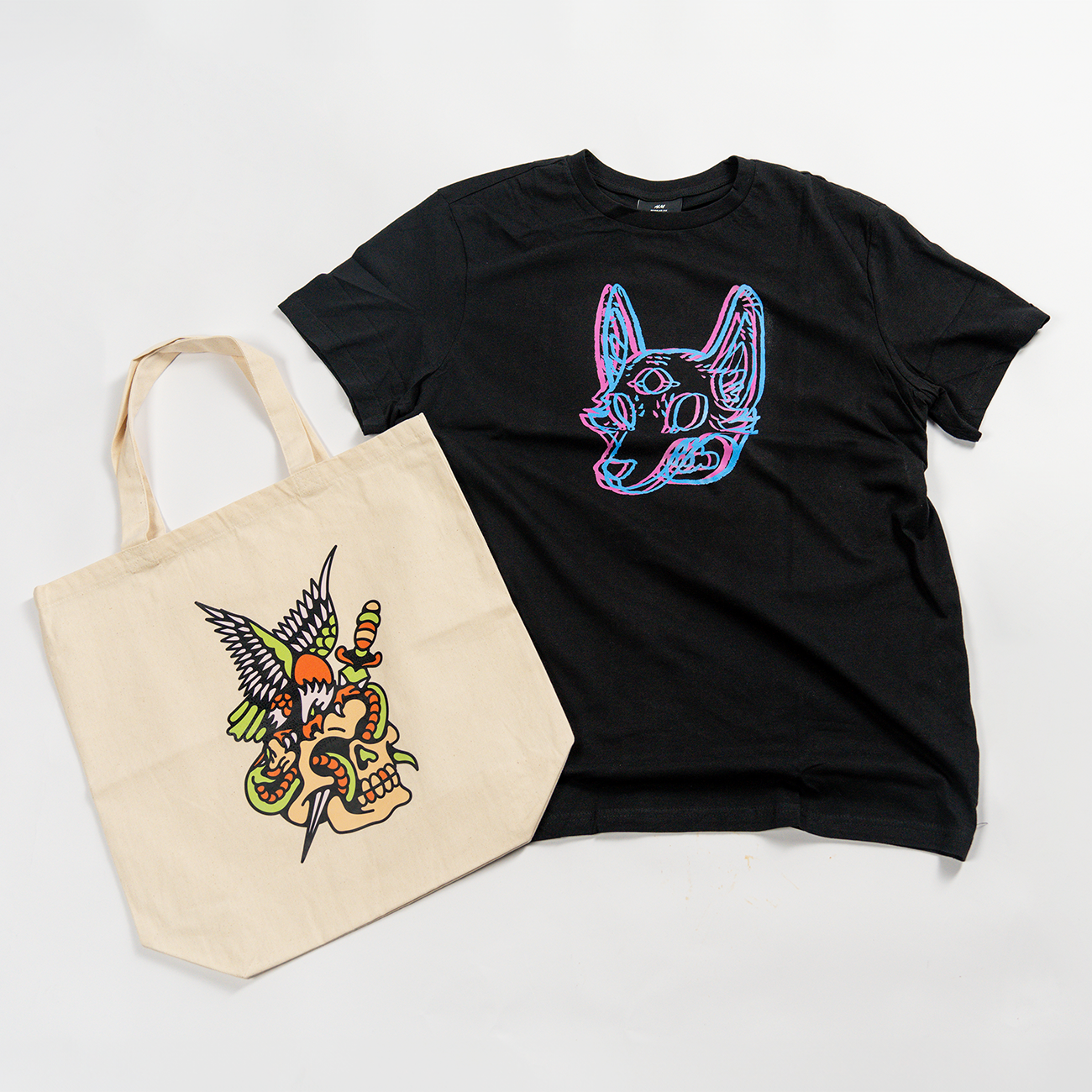 Black T-shirt with colorful heat transfer dog design and beige tote bag featuring a vibrant eagle and snake illustration with heat transfer print
