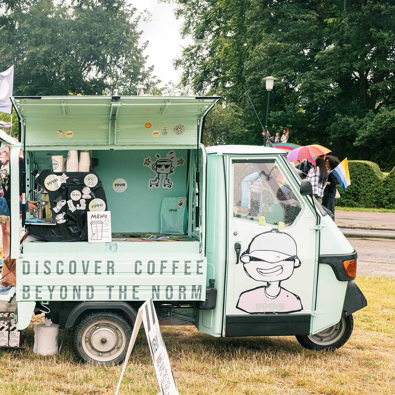 A mint-green coffee truck with stickers offers drinks at an outdoor event. People with umbrellas are in the background.