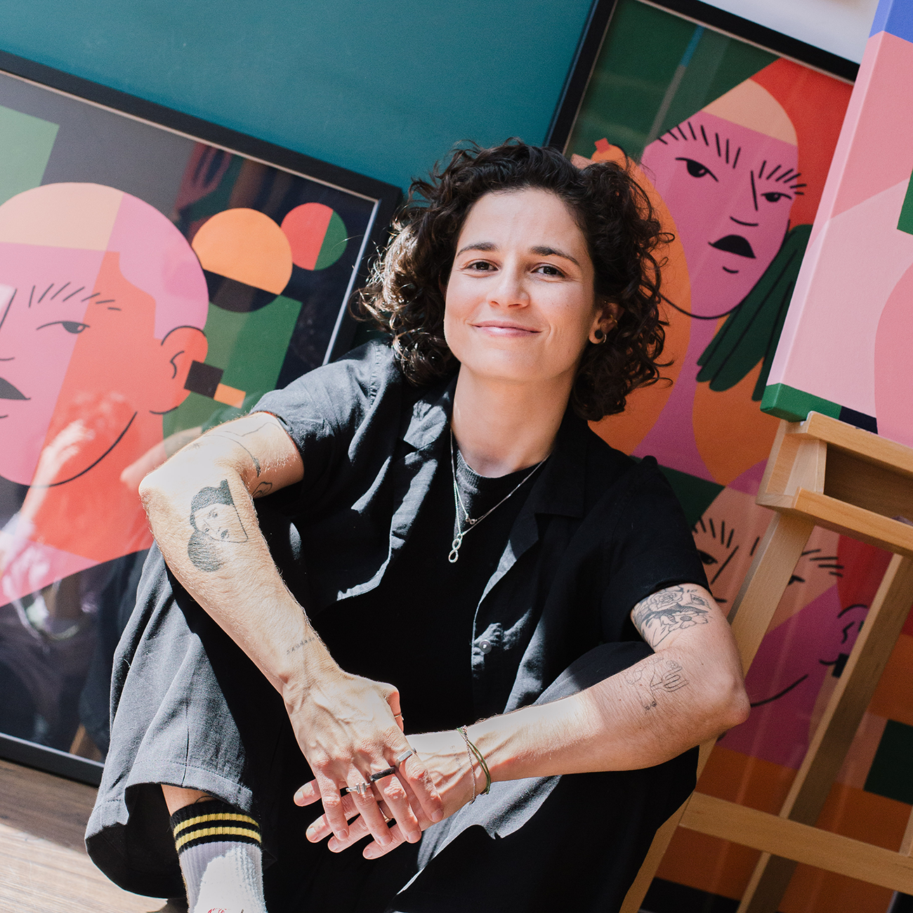 Person sitting on the floor, smiling, surrounded by colorful abstract artwork, wearing a black outfit and showing tattoos on their arms.