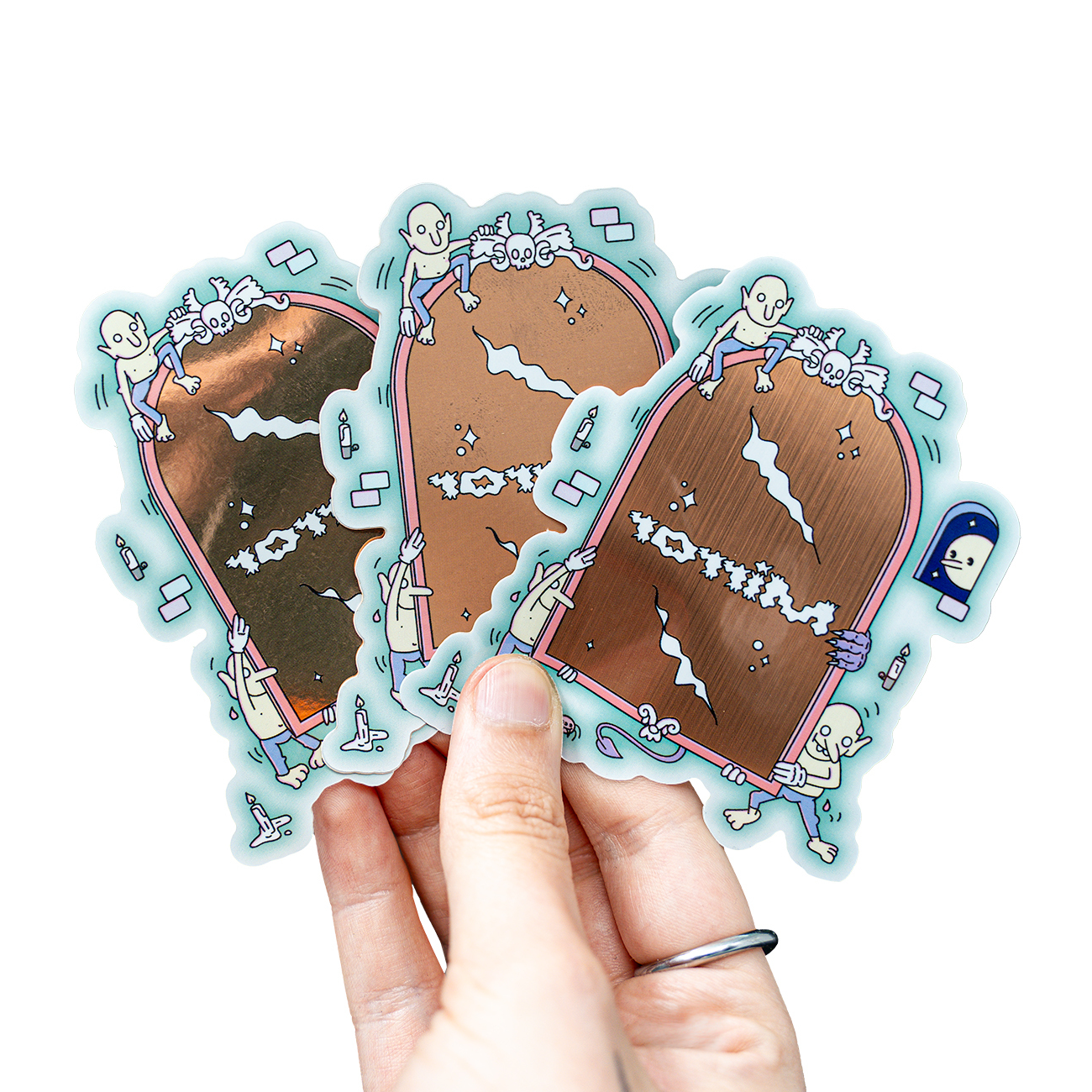 Hand holding three metallic stickers featuring cartoon skeletons climbing on rose gold mirrors with playful designs.