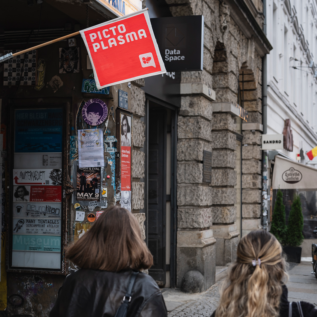 Pictoplasma flag hanging from a building, with art displayed in the background and two women walking towards it.