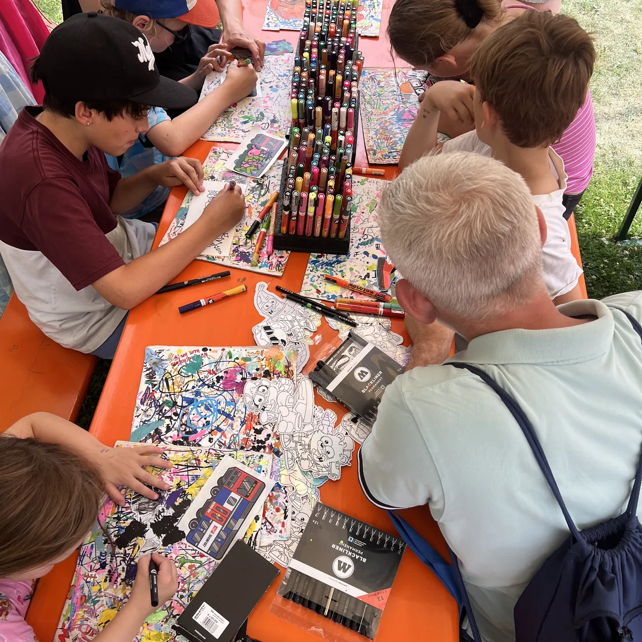 Kids colouring Brainfart die-cut stickers around a rack of paint markers at an outdoor festival workshop
