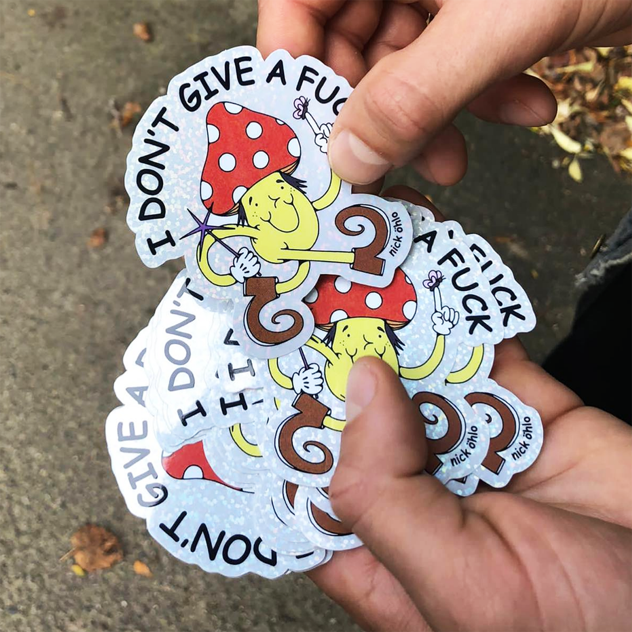 Hands holding glitter stickers of a happy mushroom cartoon man with a red hat, and quirky boots, with the text "I don't give a fuck".
