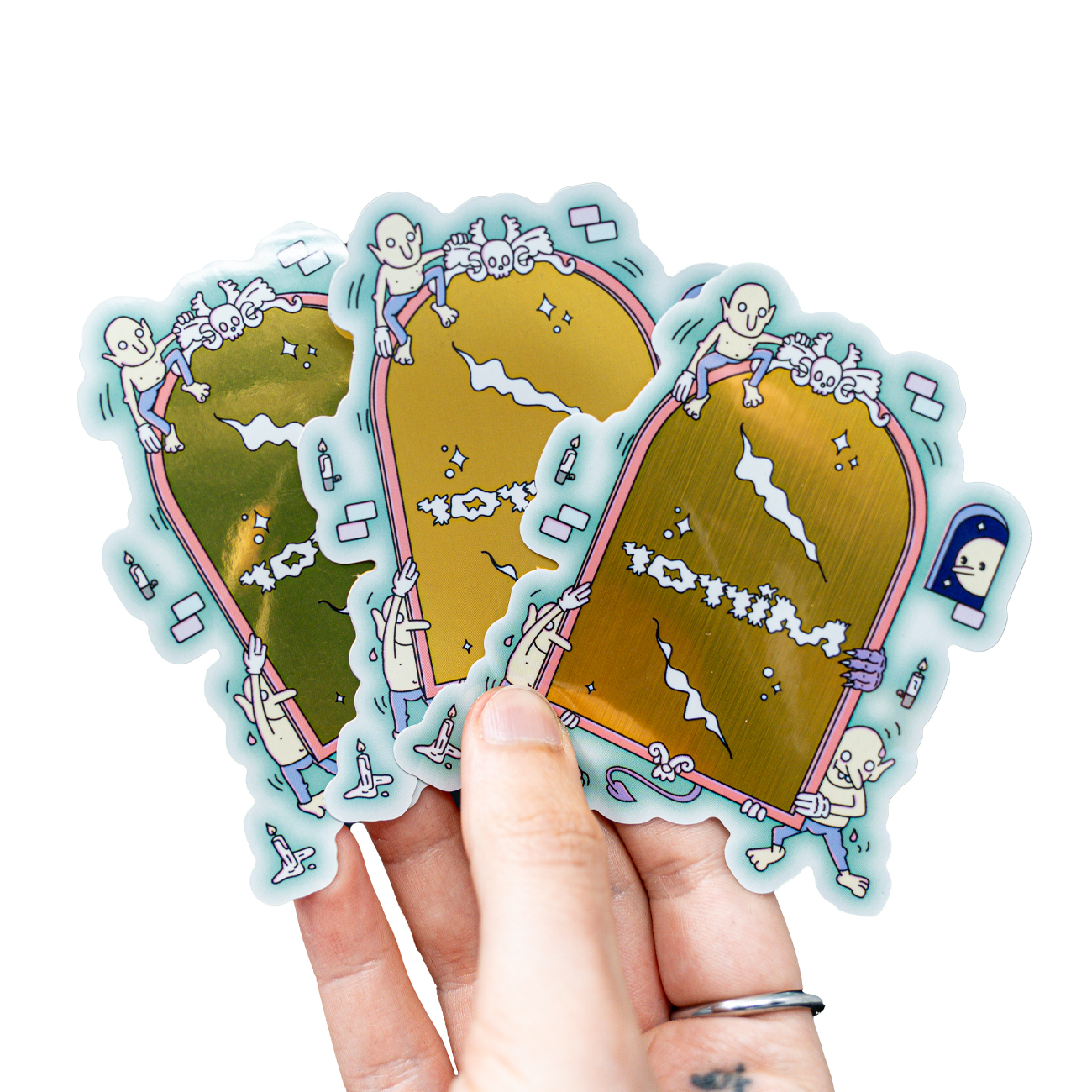 Hand holding three metallic stickers featuring cartoon skeletons climbing on golden mirrors with playful designs.