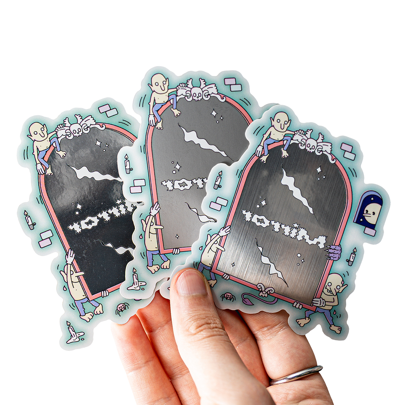Hand holding three metallic stickers featuring cartoon skeletons climbing on steel mirrors with playful designs.