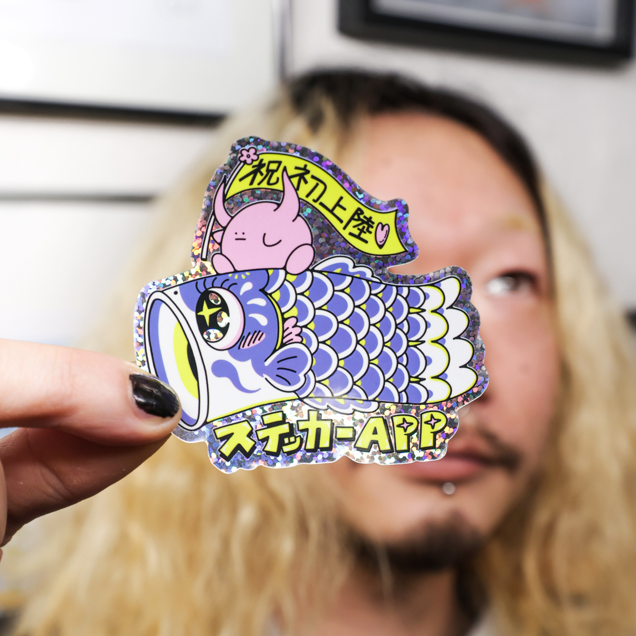 Person with blonde hair holding a shiny die-cut glitter sticker featuring a cute character on a colorful fish with Japanese text.