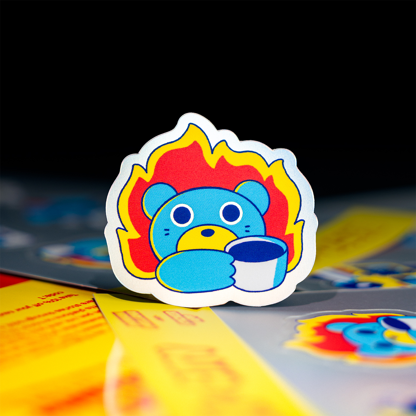 Sticker of a blue bear with a flaming background, holding a mug. The bear has a surprised expression. Bright colors and a playful design.