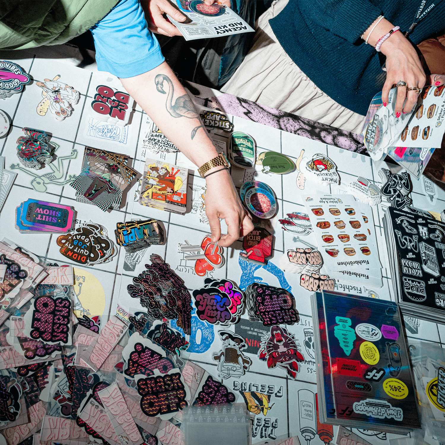 People browsing a table filled with colorful die cut stickers, sticker sheets, and sticker packs, reaching for items.