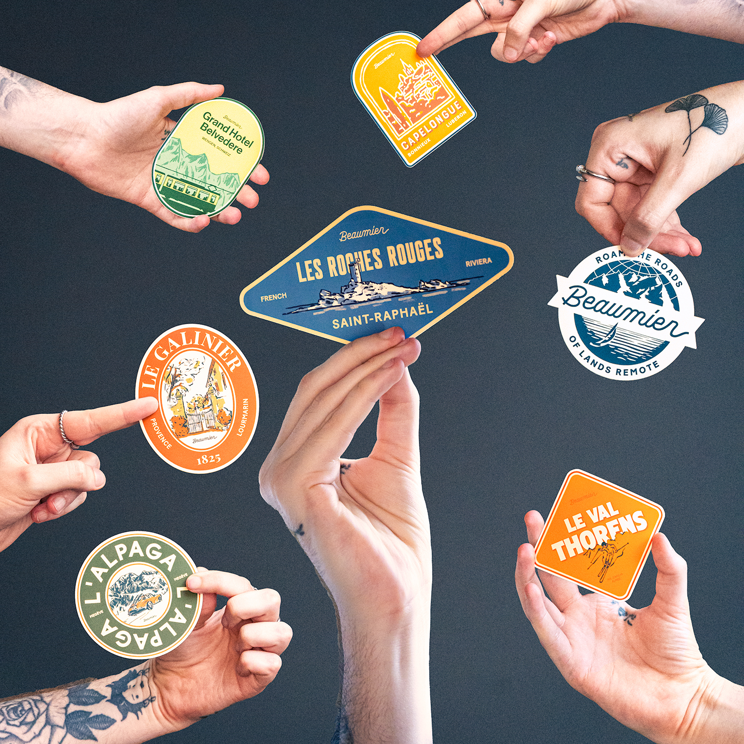 Hands holding various colorful vintage-style stickers against a dark background, featuring designs and text in French.