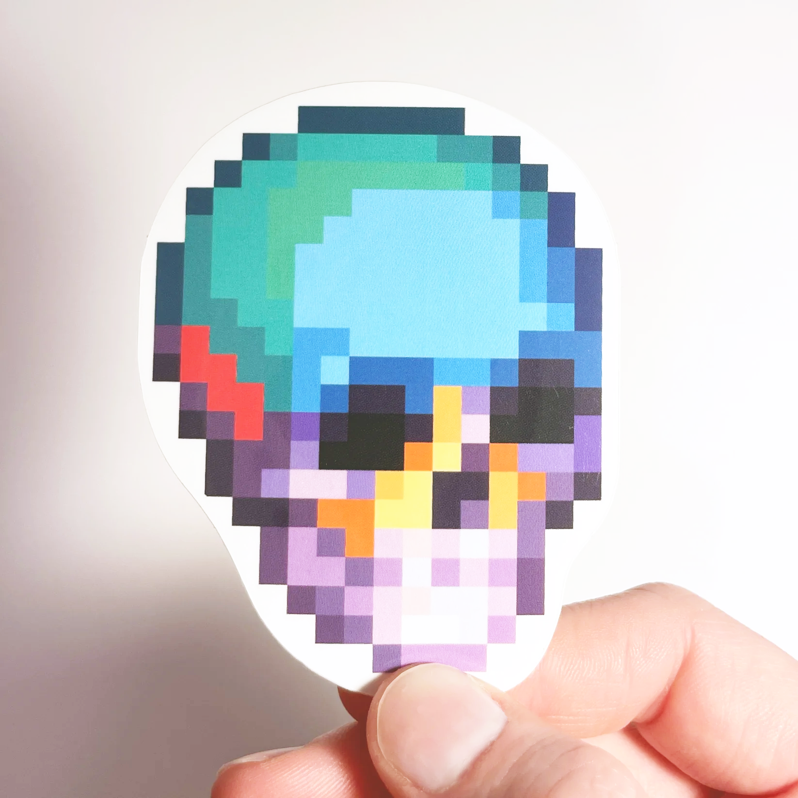 Hand holding a colorful pixel art skull sticker with shades of blue, green, purple, and orange against a plain white background.