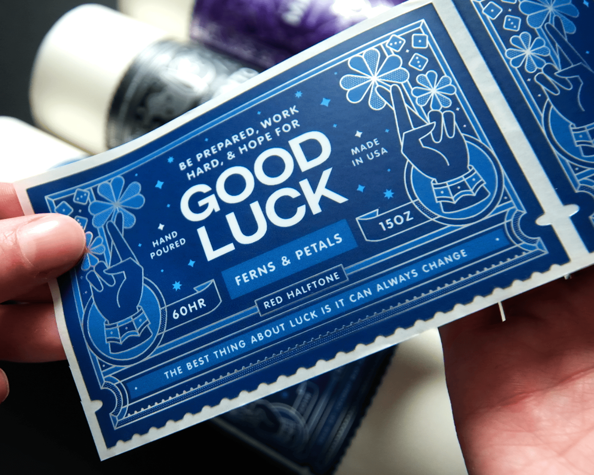 Close-up of a person holding a blue candle label featuring a hand holding a flower and the text "Good Luck" with various product details.