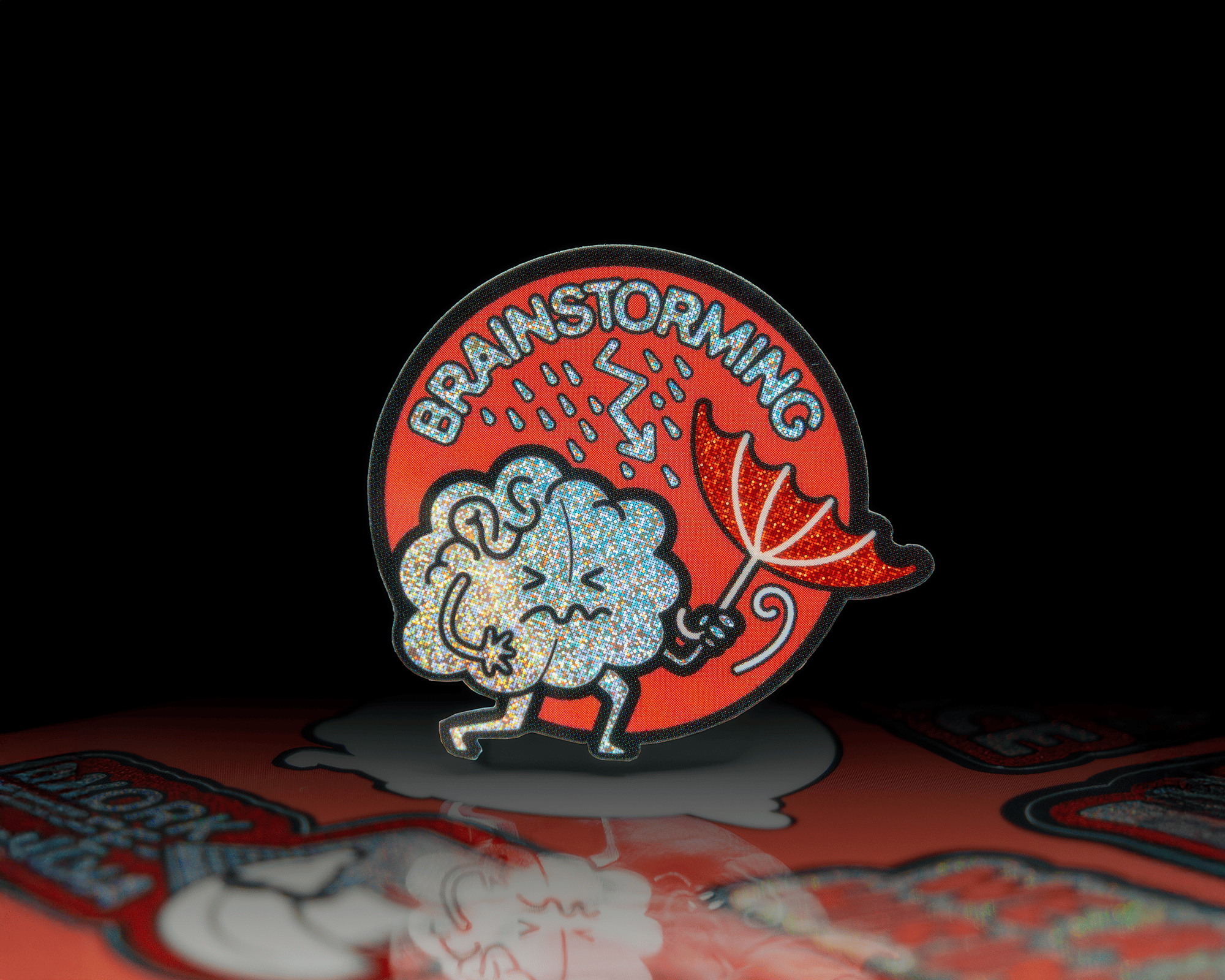 Pixie dust sticker of a brain holding an umbrella under rain, with "BRAINSTORMING" text on a red circular background.