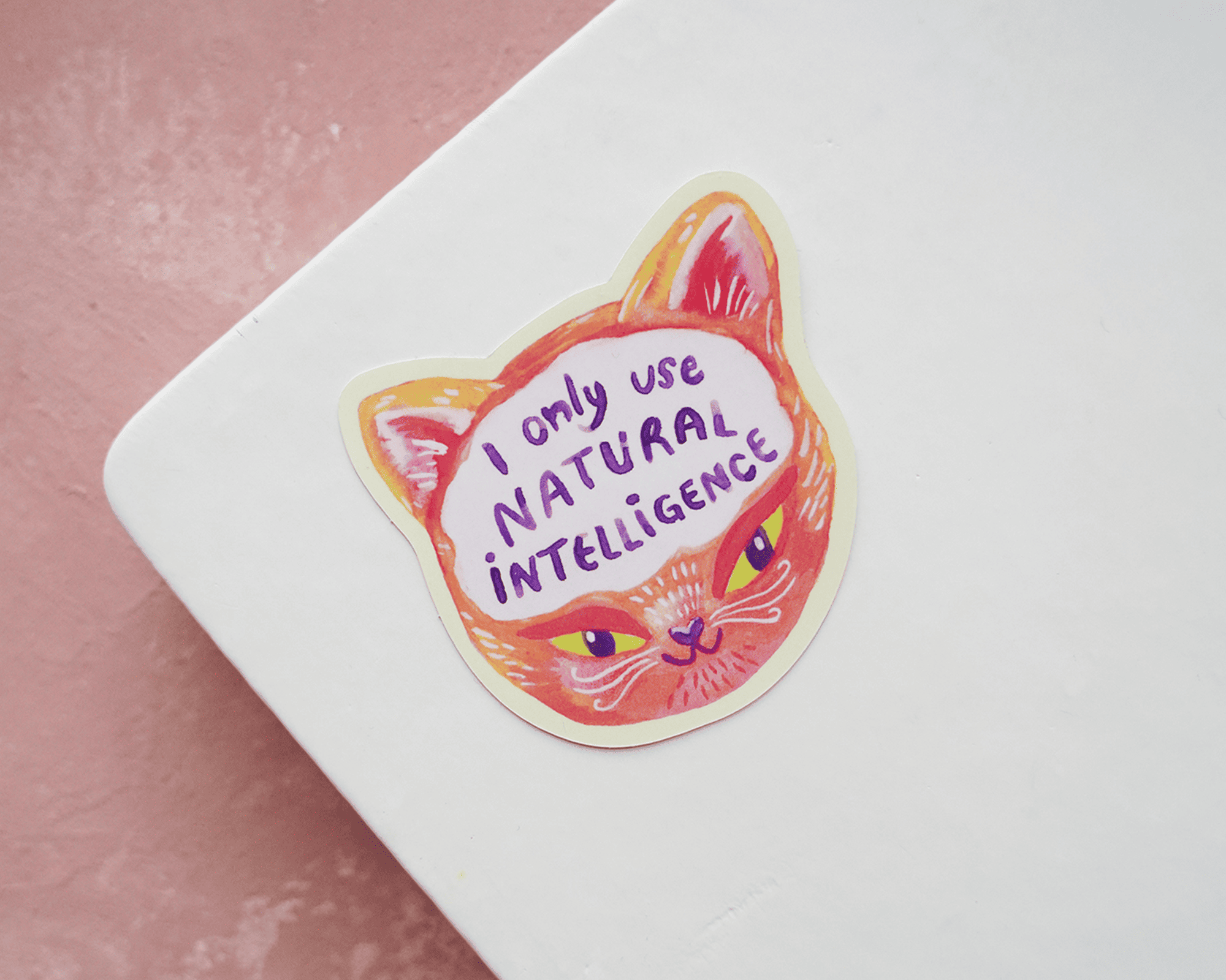 Die-cut cat face sticker reading "I only use natural intelligence" on a white notebook