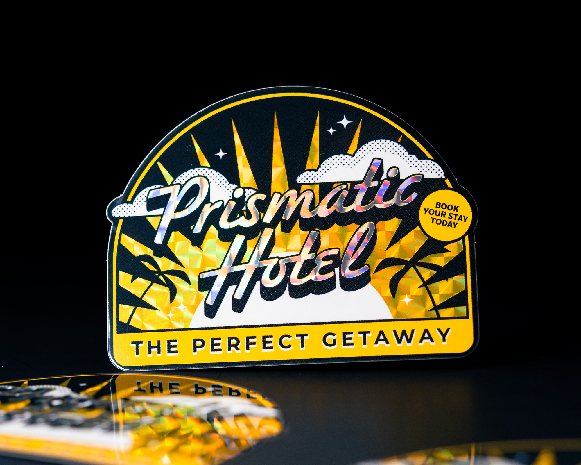 Sticker of Retro-style "Prismatic Hotel" sign with sunburst, clouds, and palm trees on a black background; reads "The Perfect Getaway" and "Book Your Stay Today."