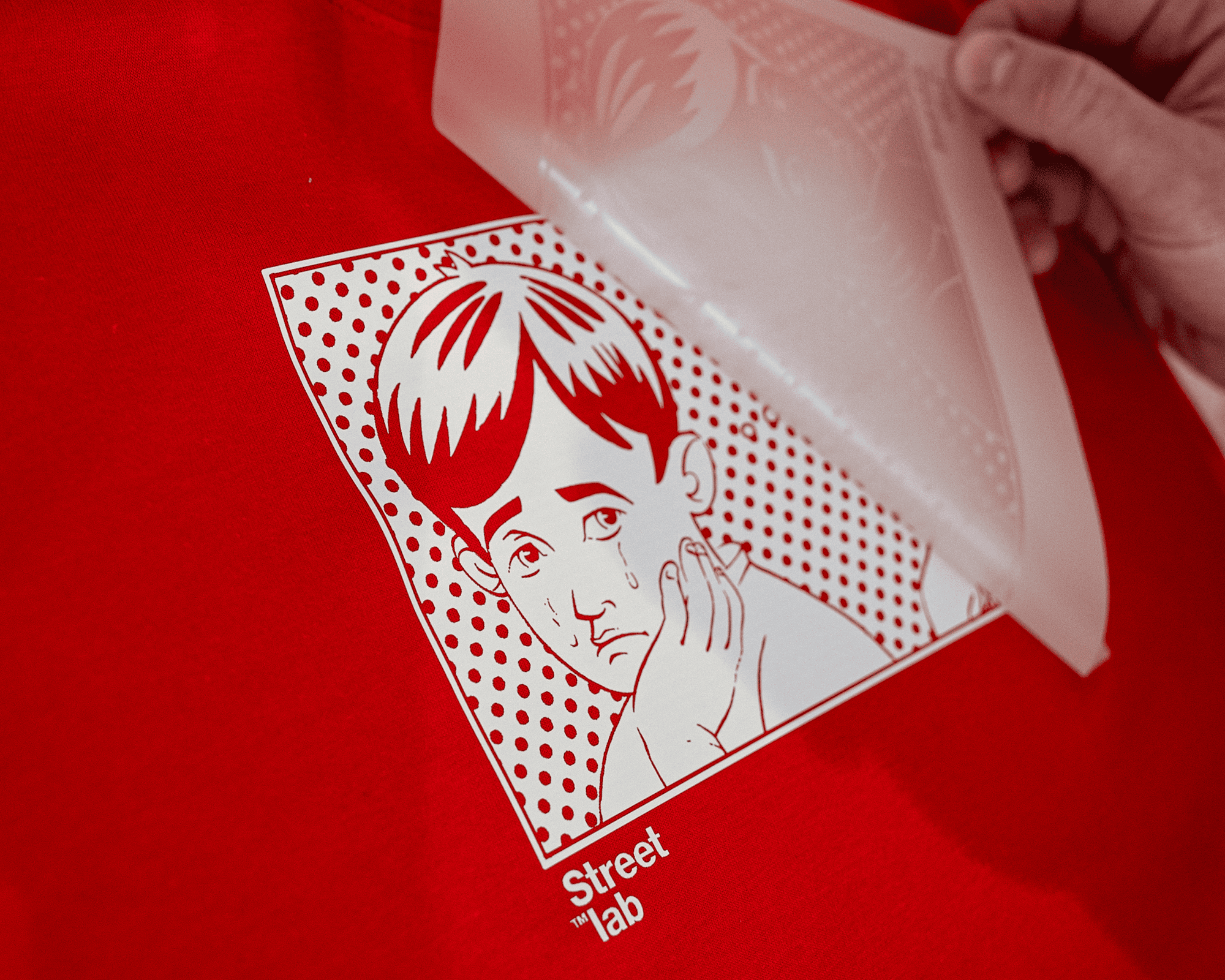 A hand peels a transfer sticker off a red surface, revealing a pop art style image of a sad boy with "Street Lab" text below.