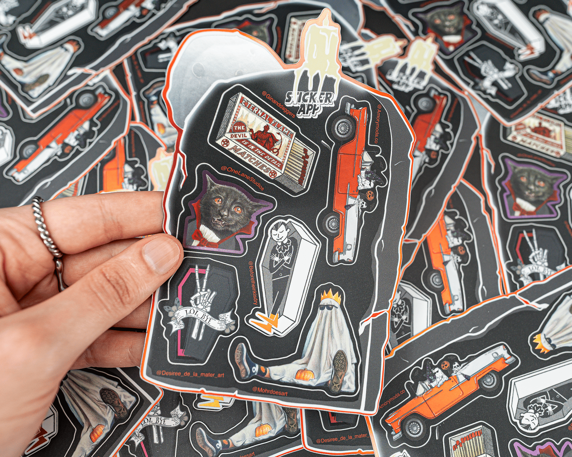 A hand holding Halloween-themed sticker sheet, including a cat, coffin, vampire, ghost, and hearse, with more stickers in the background.