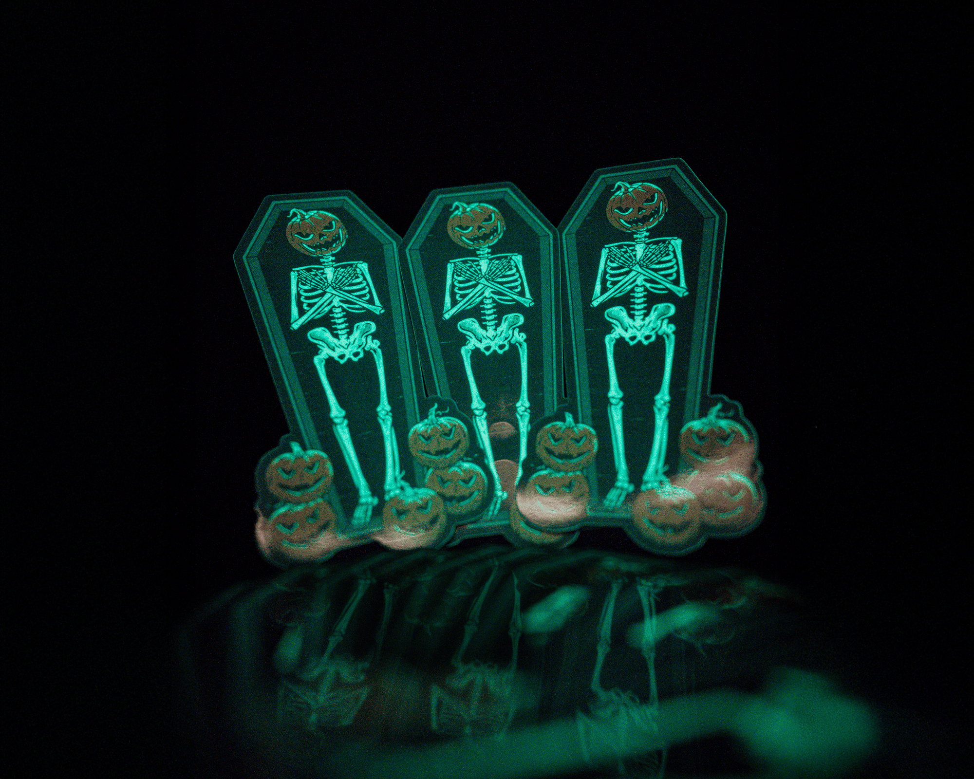 Three glow in the dark skeleton stickers with pumpkin heads stand in a row, surrounded by small pumpkins, against a dark background.