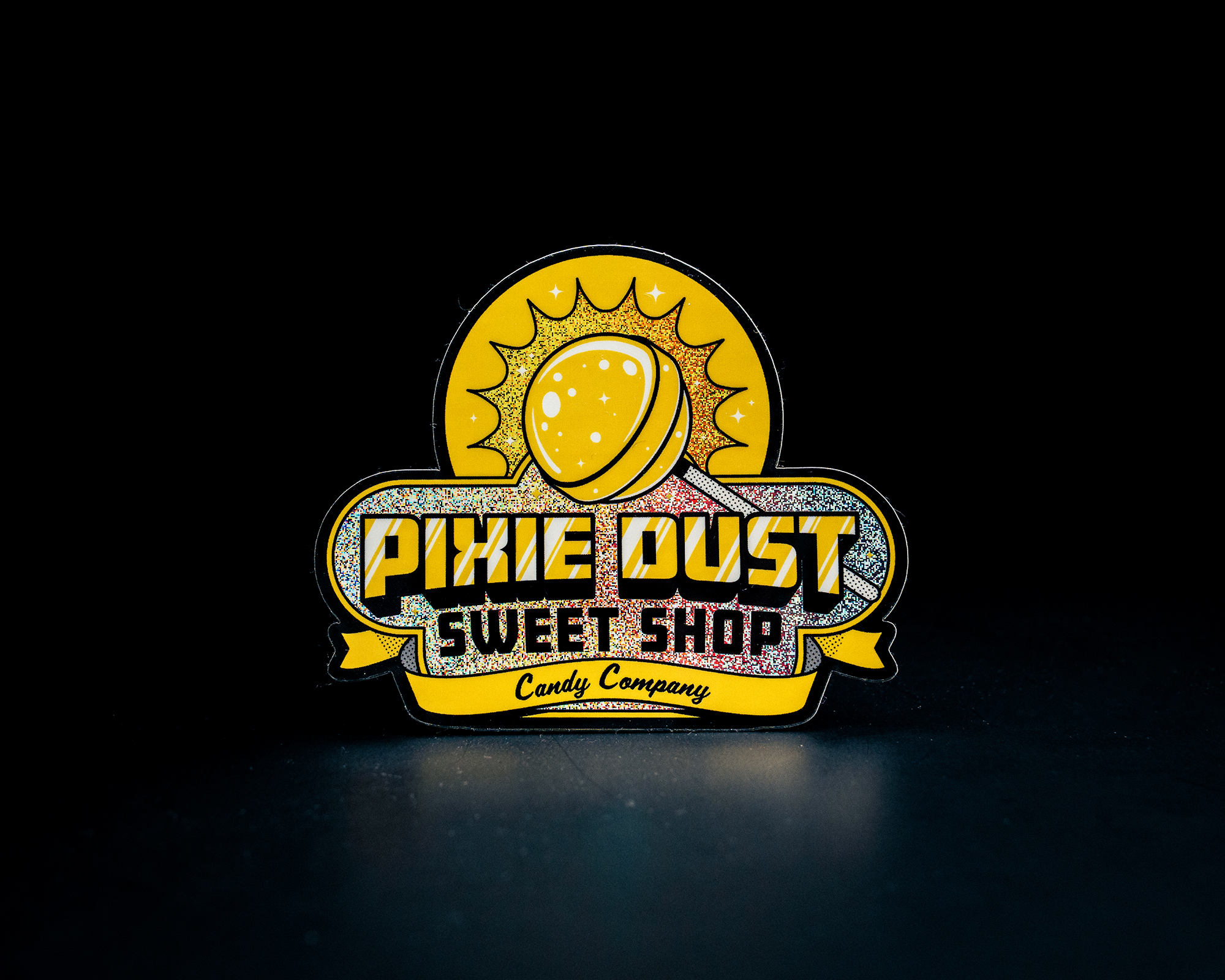 Yellow and glittery Pixie Dust Sweet Shop sticker featuring a lollipop design on a dark background.