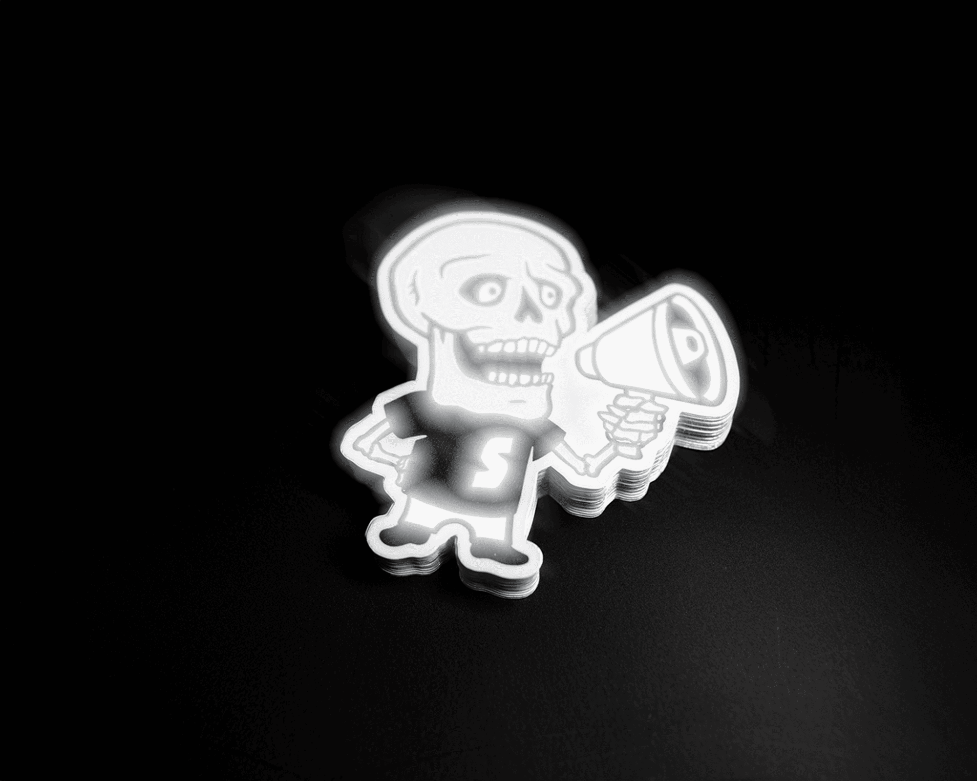 Reflective sticker of Cartoon skeleton with a megaphone, wearing a shirt with an "S" on it, against a dark background.