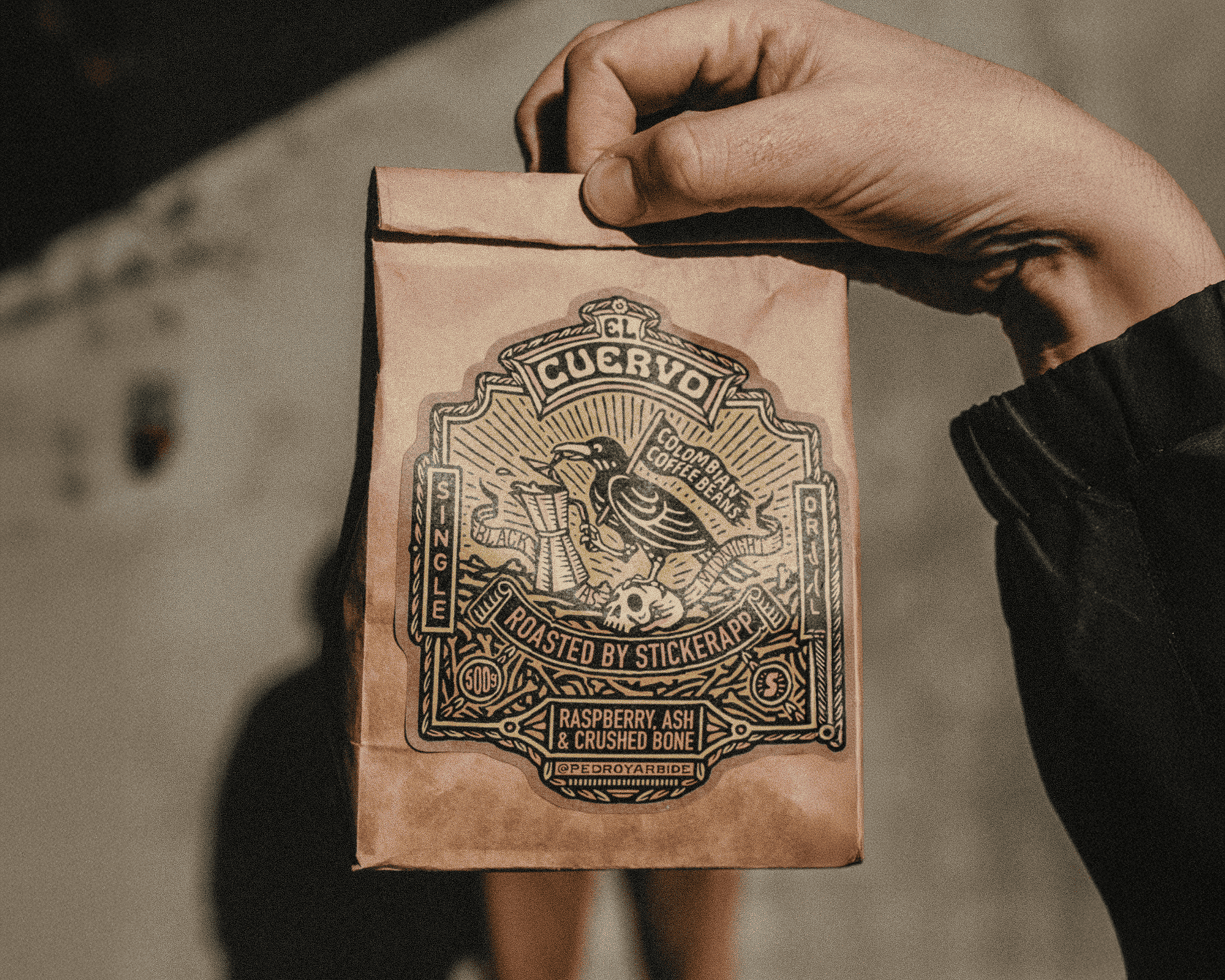Hand holding a brown coffee bag with an ornate kraft label featuring a skeleton and text about the coffee blend, "Raspberry Ash & Crushed Bone."