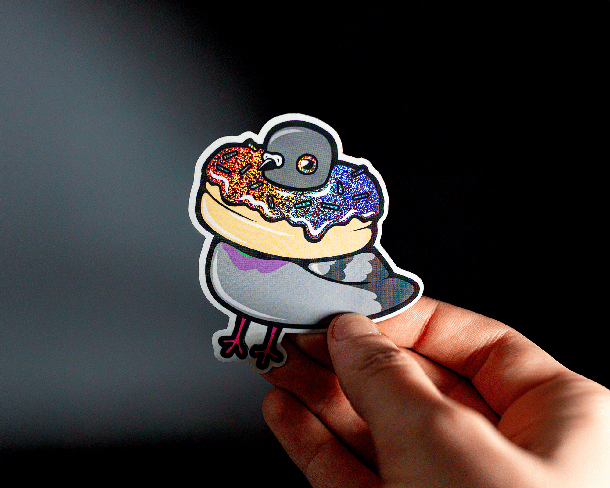 Hand holding a pixie dust sticker of a cartoon pigeon wearing a colorful donut around its neck, set against a dark background.