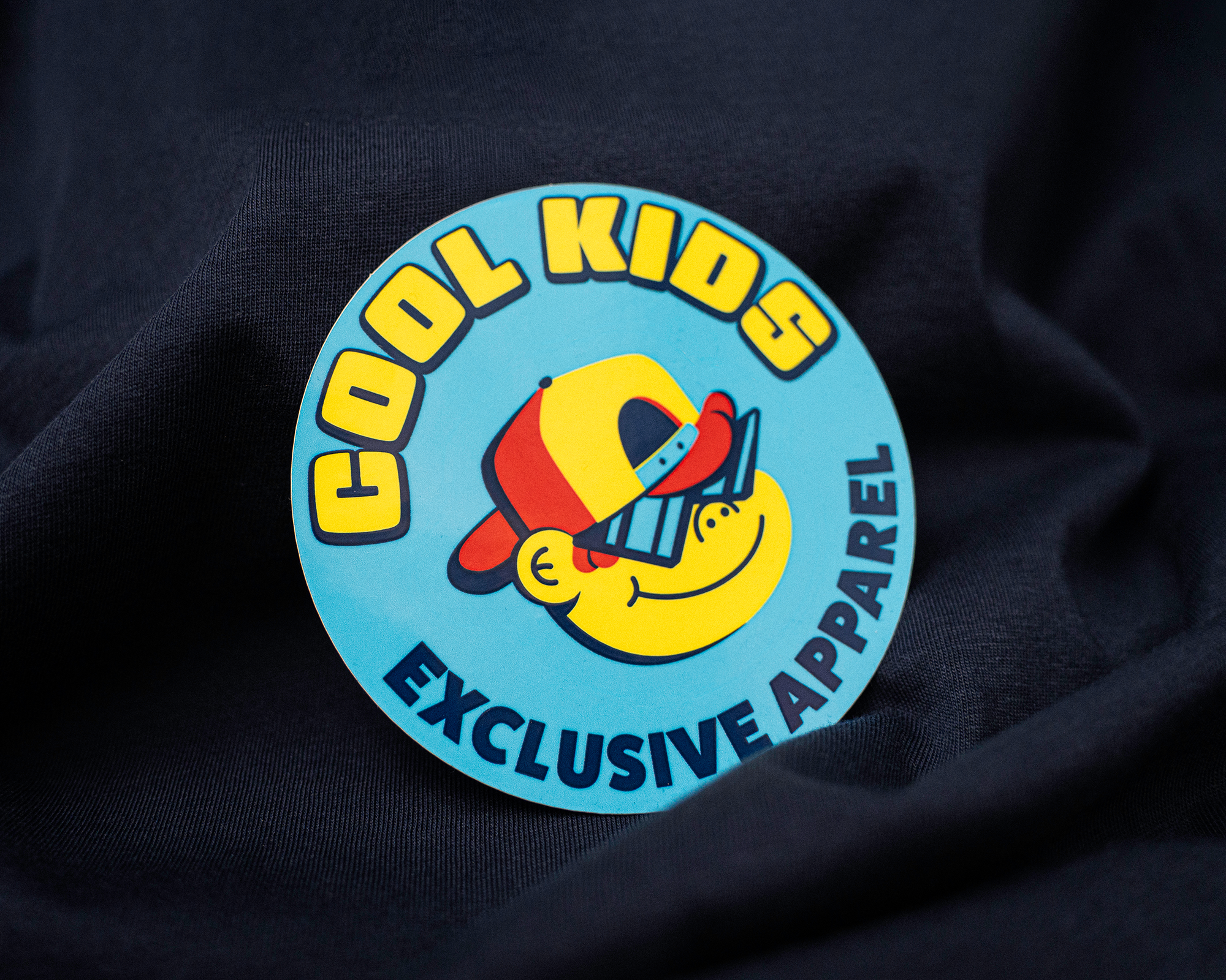 Round blue sticker featuring a cartoon child wearing a sideways red-and-yellow cap and oversized sunglasses.