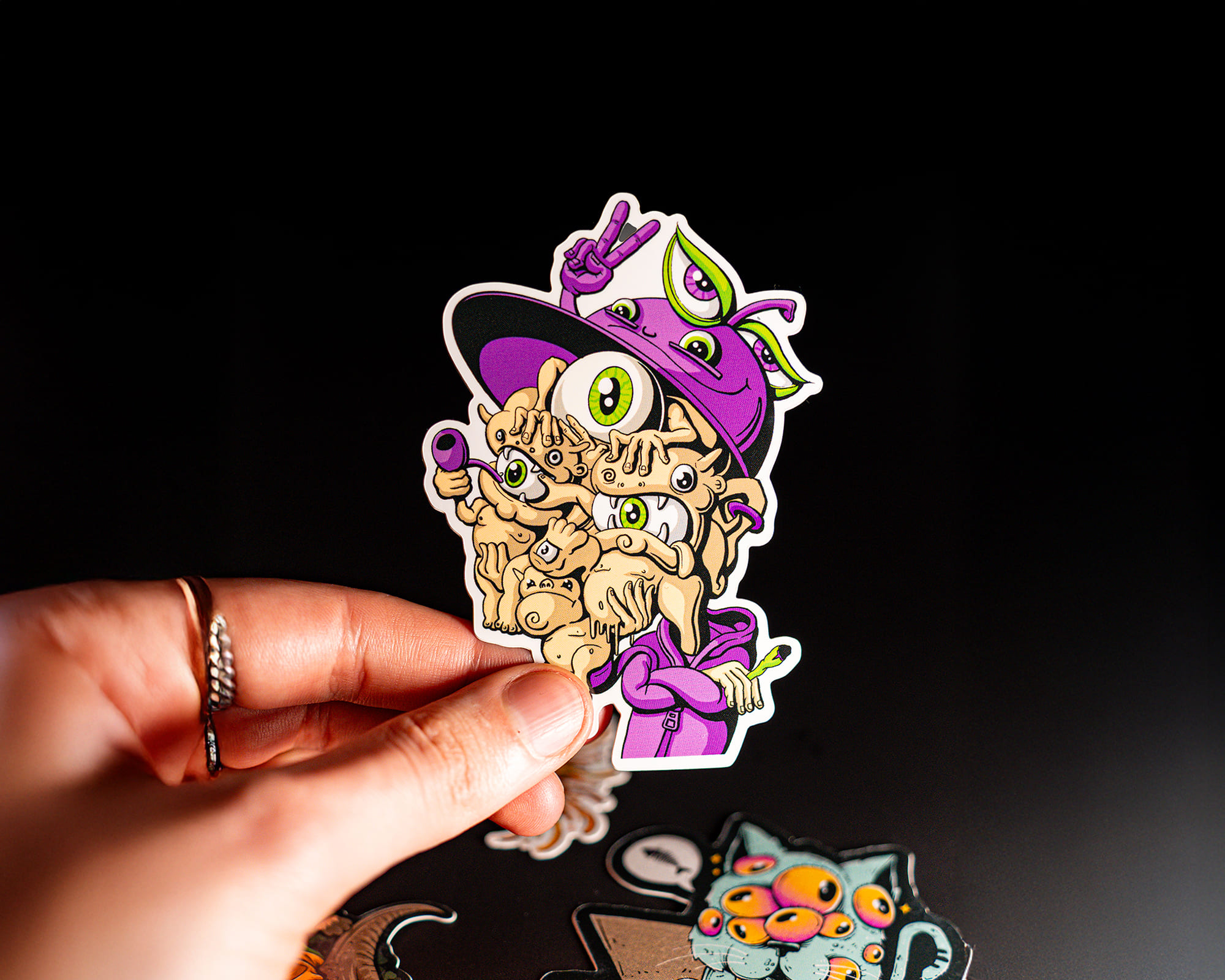 Hand holding a colorful vinyl sticker of a cartoon character with multiple eyes, wearing a hat and making a peace sign.