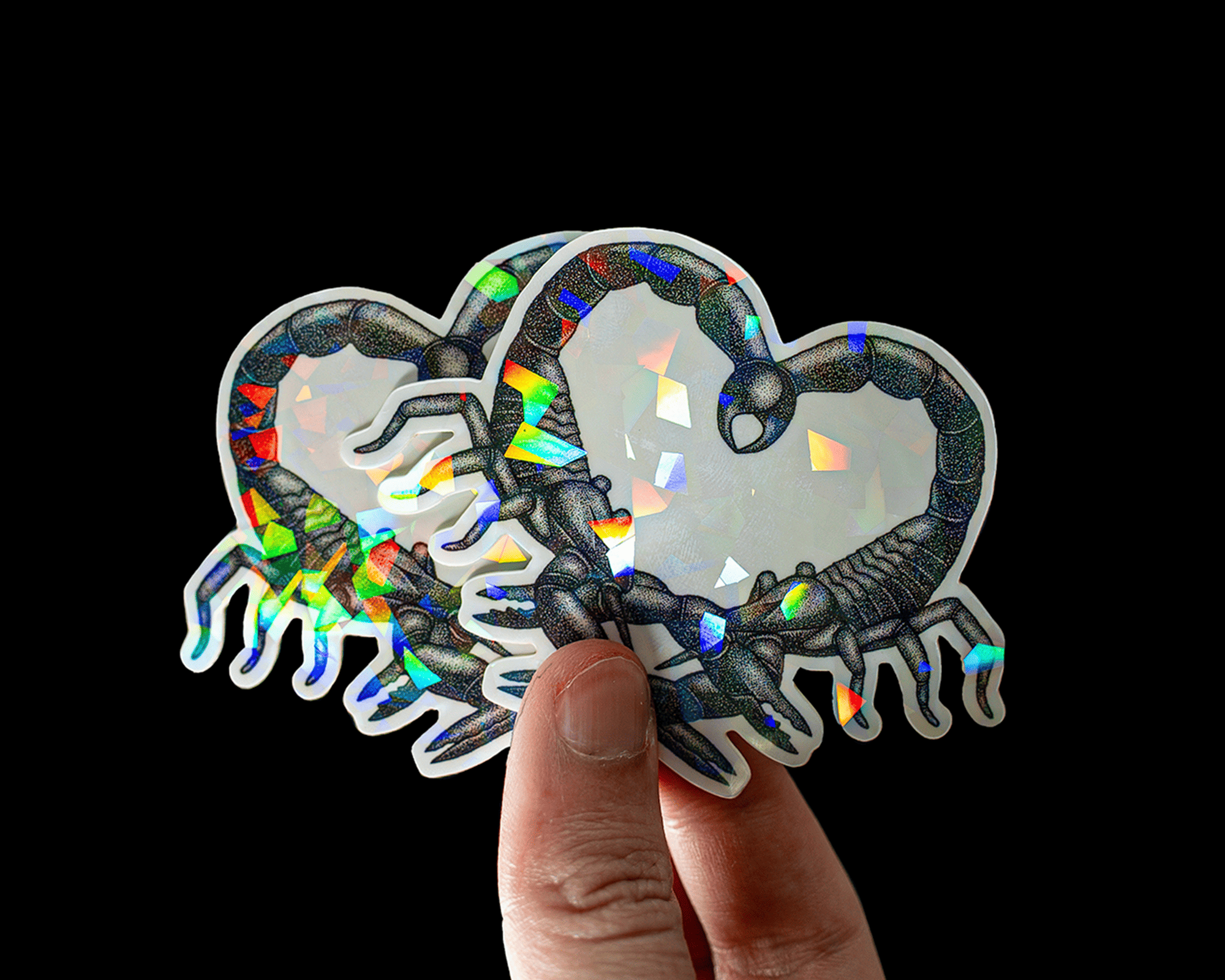 Hand holding two holographic cracked ice heart-shaped stickers with scorpion designs on a black background.