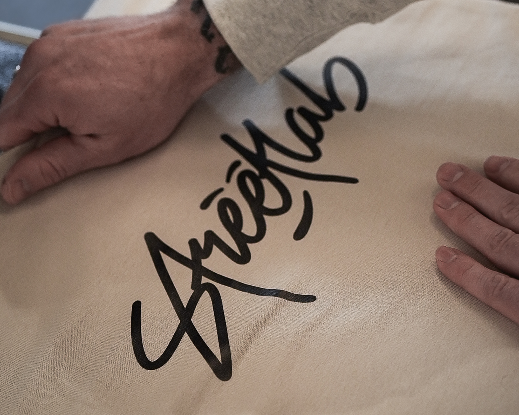 Two hands rest on a fabric with bold, stylized black text. The left hand has a tattoo on the wrist.