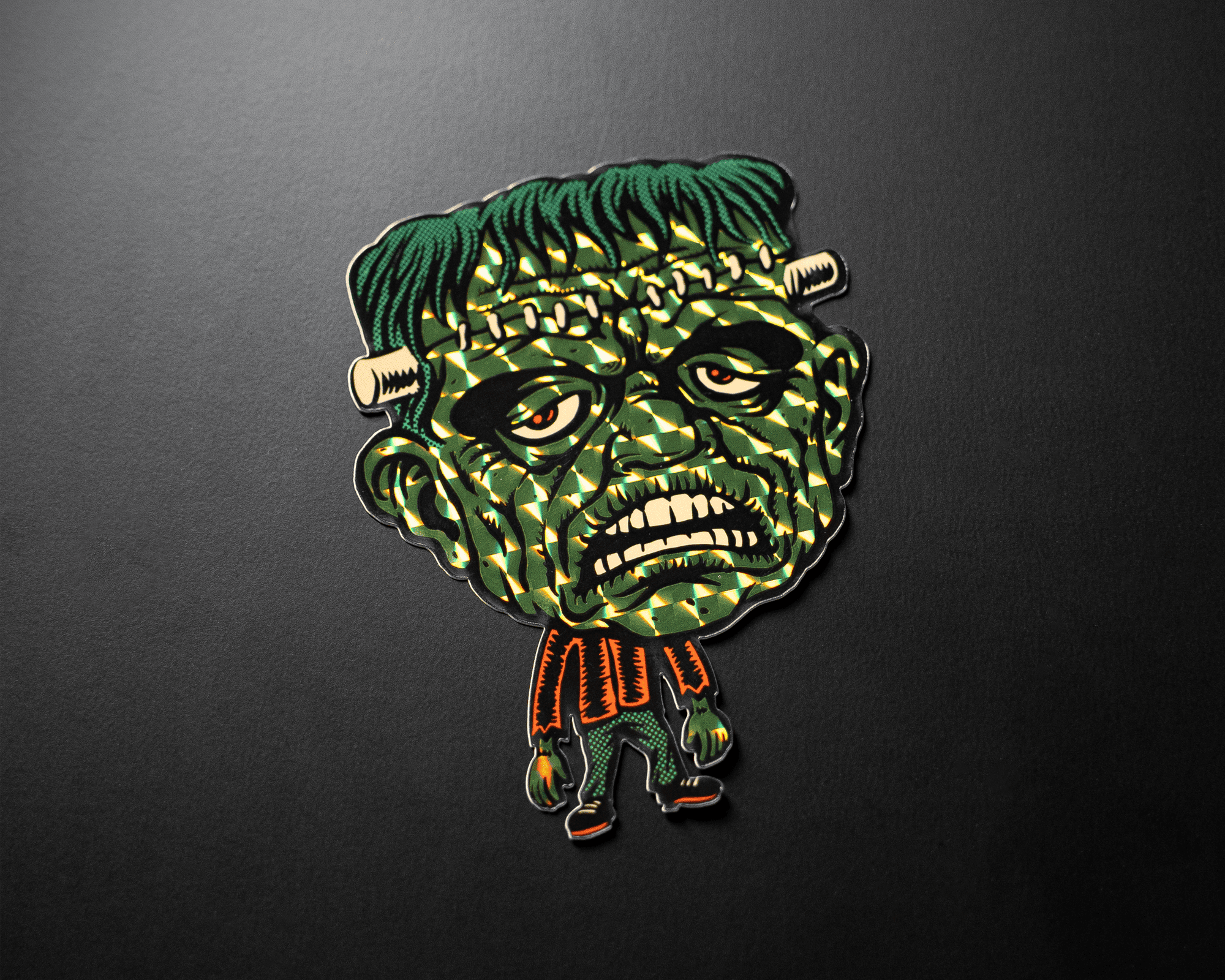 Cartoonish Frankenstein monster prismatic sticker with green skin, bolts in neck, and orange shirt on a dark background.