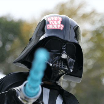 Person in a Darth Vader costume with "Who's Your Daddy?" on the helmet, holding a glowing blue lightsaber.