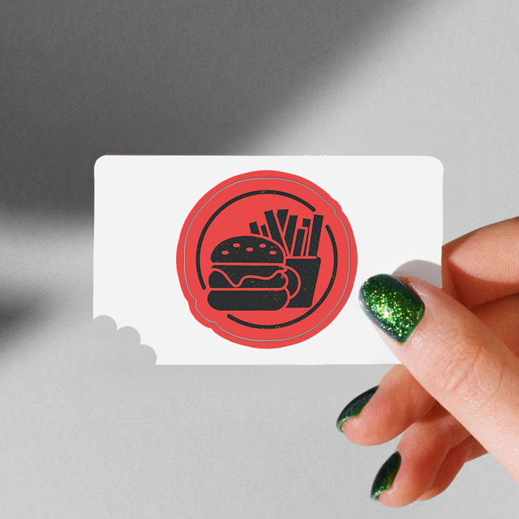 Hand with green glitter nails holding a business sticker card featuring a red circle with a burger and fries icon.