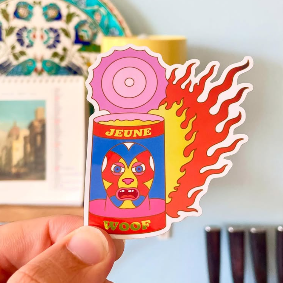 Sticker of a colorful, abstract face on a can with flames and the words "Jeune Woof." Background shows blurred decorative plates.