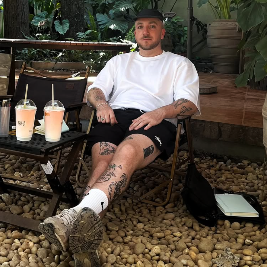 Tattoo artist and illustrator Bambi Bakbi sitting casually in a chair, with visible tattoos and drinks on a table beside him.