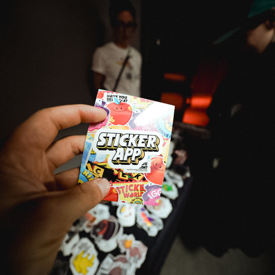 Hand holding a kiss-cut sticker with the StickerApp logo and colorful art on the front, with a person and stickers on a table in the background.